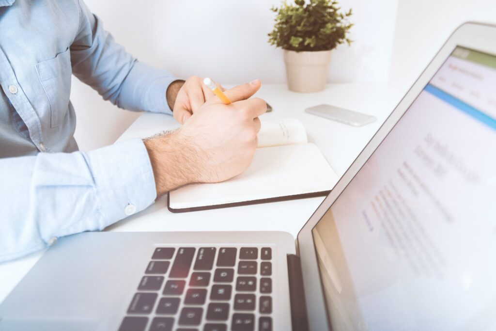 Person's hands writing in a notebook while sitting next to their Apple laptop