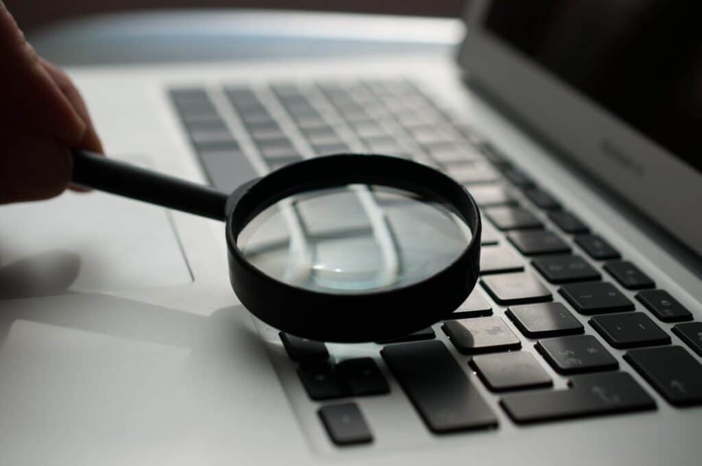 A person's hands holding a magnifying glass over a laptop keyboard