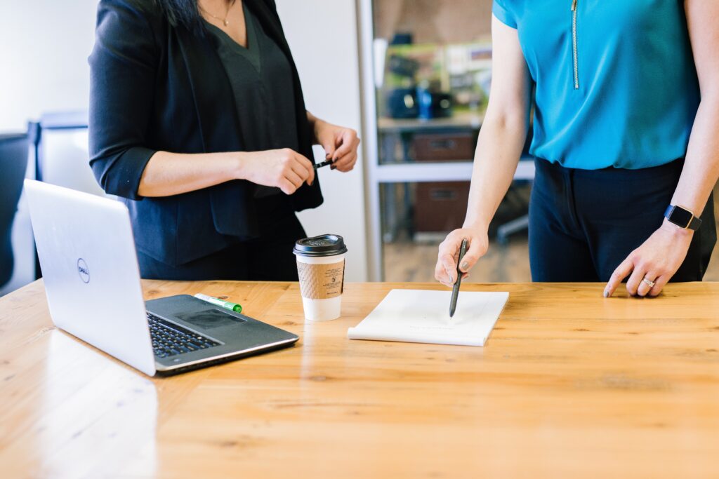 Two women from the neck down standing over a notepad, a coffee cup and a Dell laptop