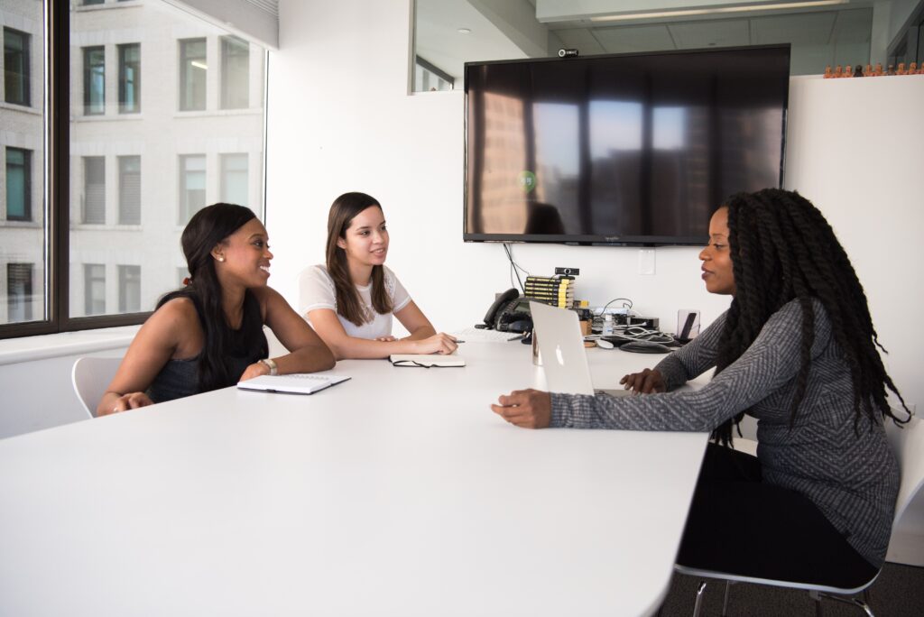 One woman with a laptop talking with two other women at a long white table in an office