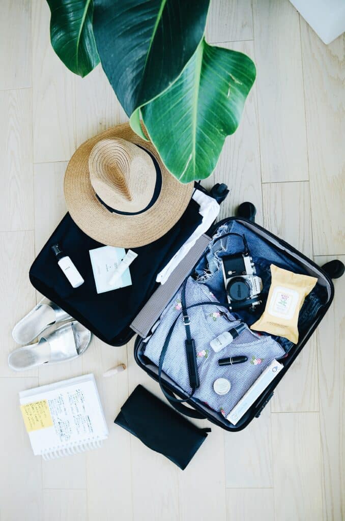 A packed open suitcase showing a camera, hat, and other items on a wooden floor with shoes, a plant and a notebook