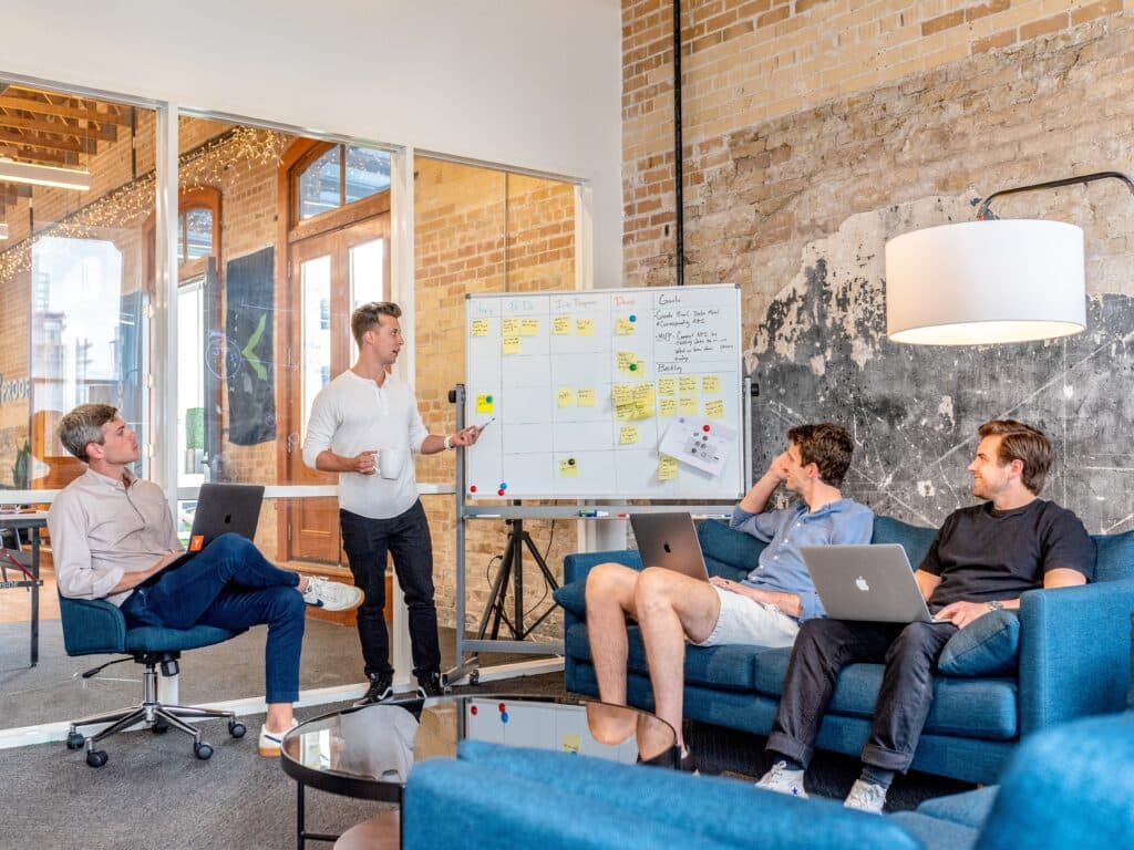 One man standing at a dry erase board talking to 3 other men on couches and chairs in a brick office meeting room