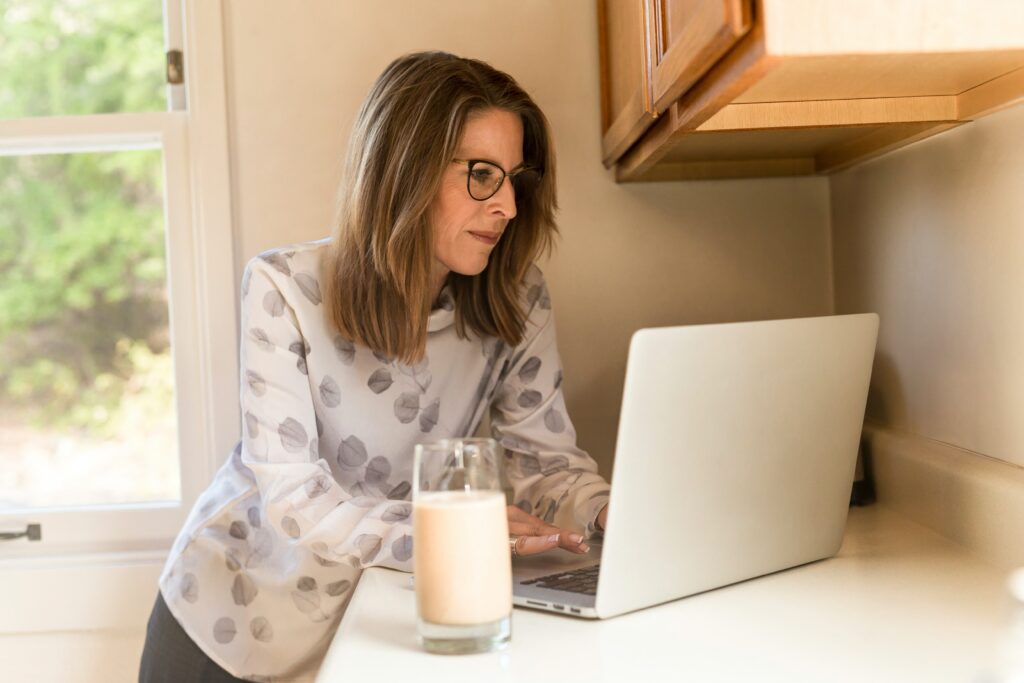 Woman with glasses leaning over a countertop working on a laptop next to a full glass