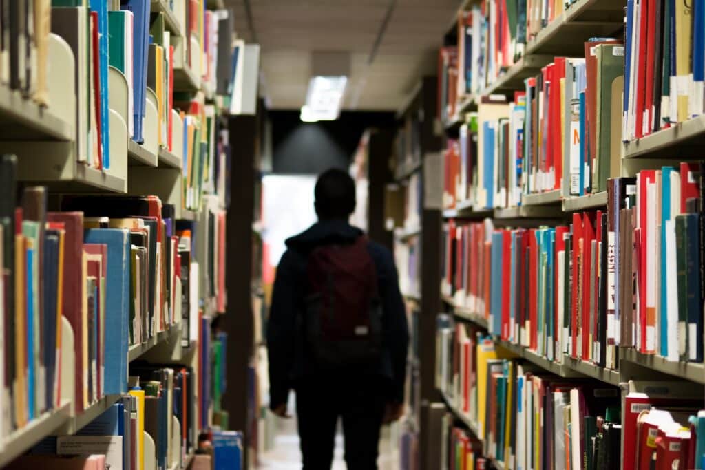 Back of a person walking with a backpack through a row of books at a library