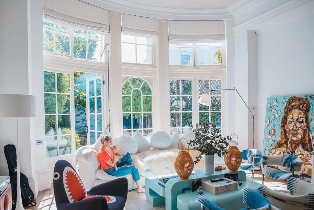 Woman sitting on a couch with a cup of coffee in a large white living room with colorful accents and large windows