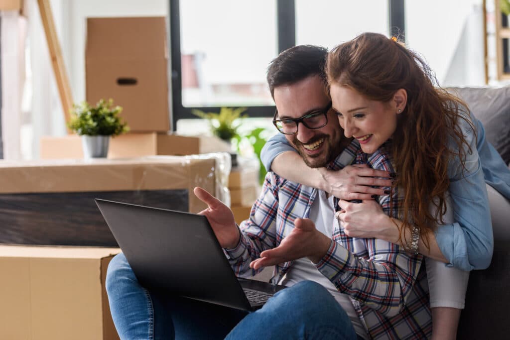 Young couple moving in new home. Sitting and relaxing after unpacking. Searching home decorating ideas on laptop