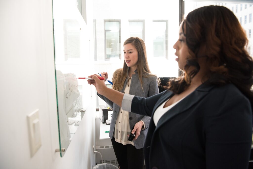 Two women standing in front of a whiteboard each with a marker