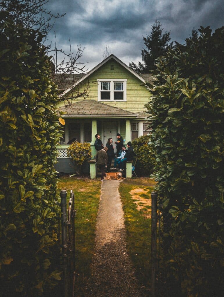 Group of people standing in front of a green house surrounding by bushes and a fence