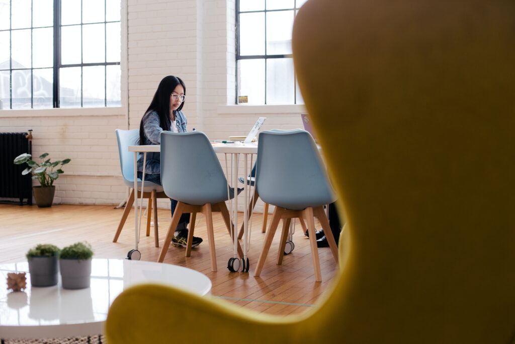 Photo taken from behind an arm chair of a woman working at a long table in a white brick office