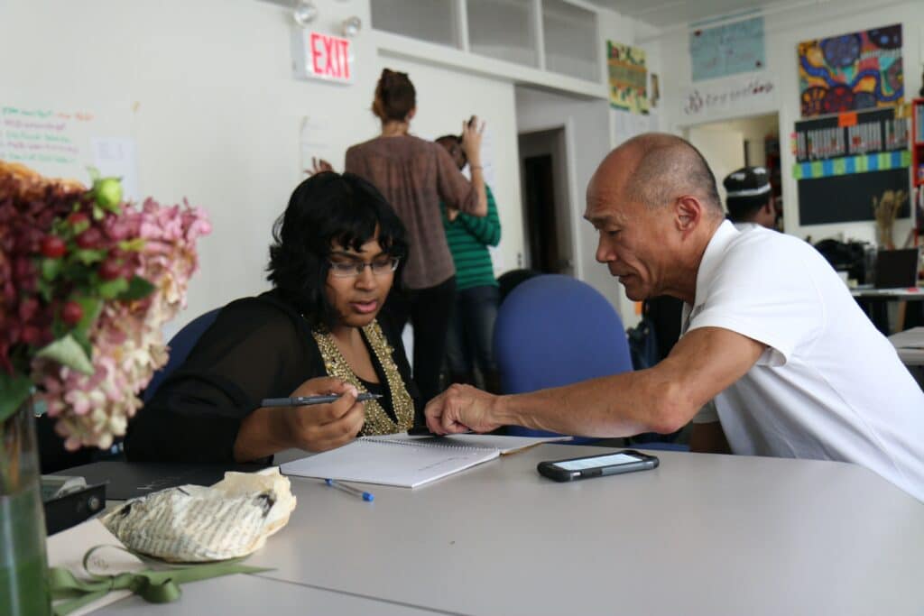 Man helping to explain something in a notebook to a woman in classroom setting