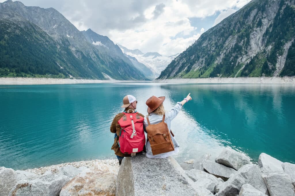 Two travelers with backpacks sit by a scenic mountain lake, planning their next adventure. Travel insurance can provide peace of mind, but is travel insurance worth it for your trip?