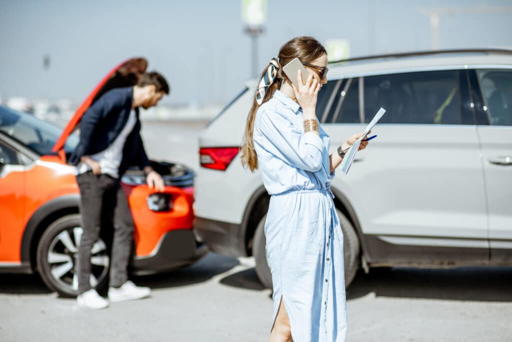 A woman on the phone reviewing paperwork after a car accident, while a man checks vehicle damage. Finding affordable car insurance can help cover unexpected incidents.