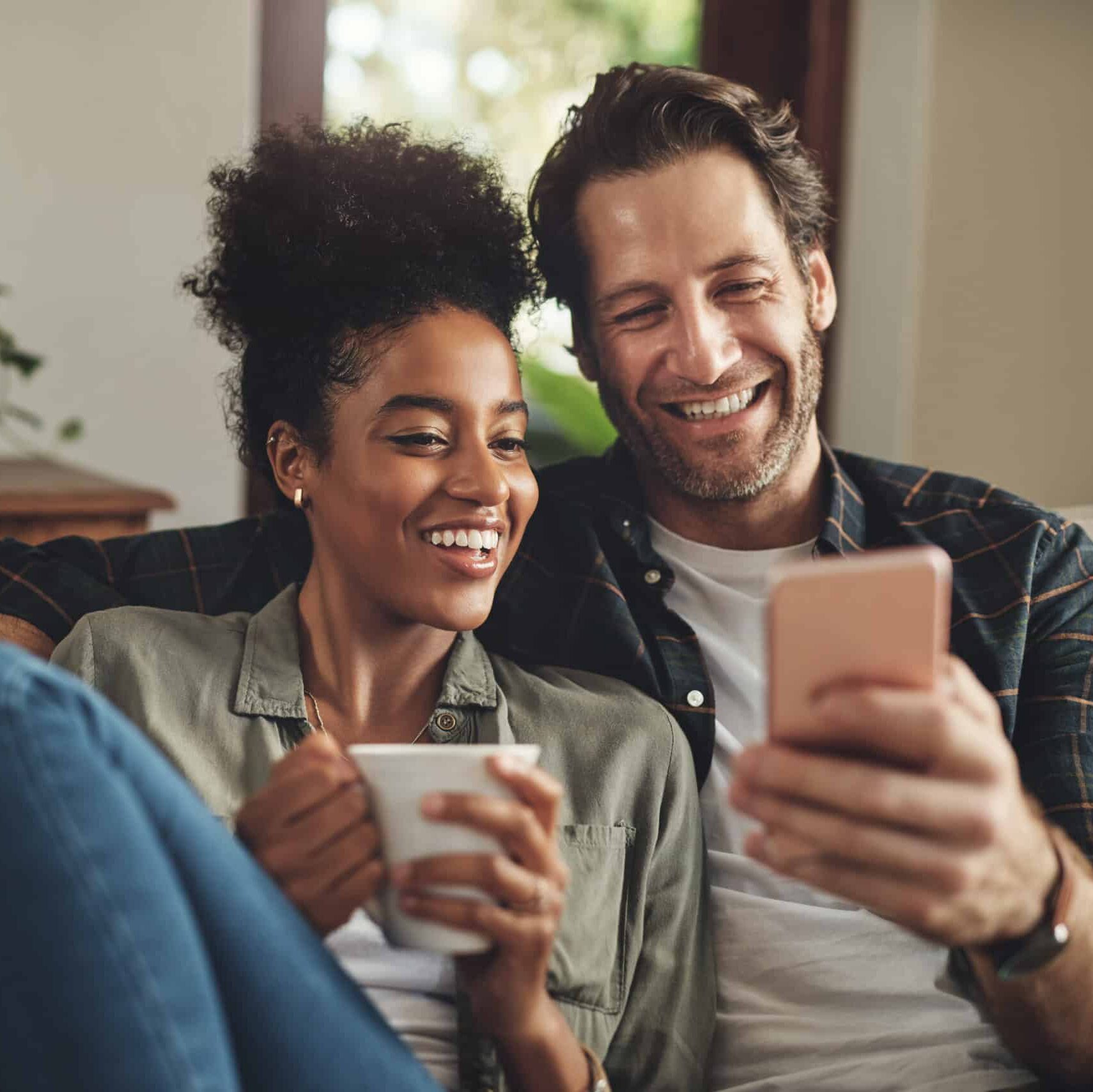 a happy young couple using a cellphone together while relaxing on a couch at home