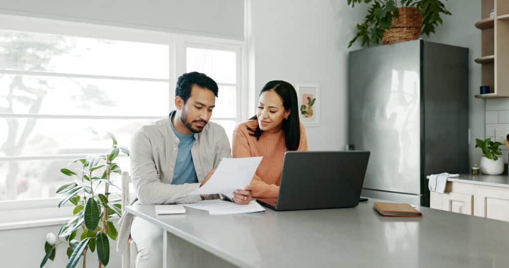 A middle-aged couple sits at their kitchen counter, reviewing financial documents together with a laptop open in front of them, possibly discussing reverse mortgage options for their home.